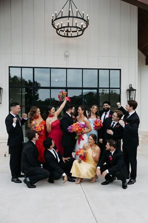 Wedding party celebrates outdoors under chandelier, bridesmaids in colorful gowns, groomsmen in black suits, holding vibrant bouquets.