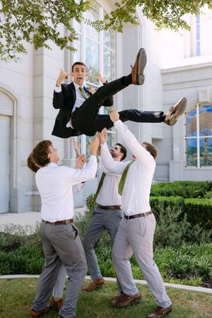 Groomsmen tossing groom in the air outside a wedding venue, all wearing suits and dress shirts.