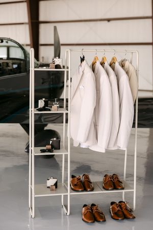 White suits on a clothing rack with brown dress shoes below, displayed in an airplane hangar setting.