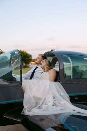 Bride and groom embrace romantically in a small aircraft cockpit, wearing wedding attire at sunset.