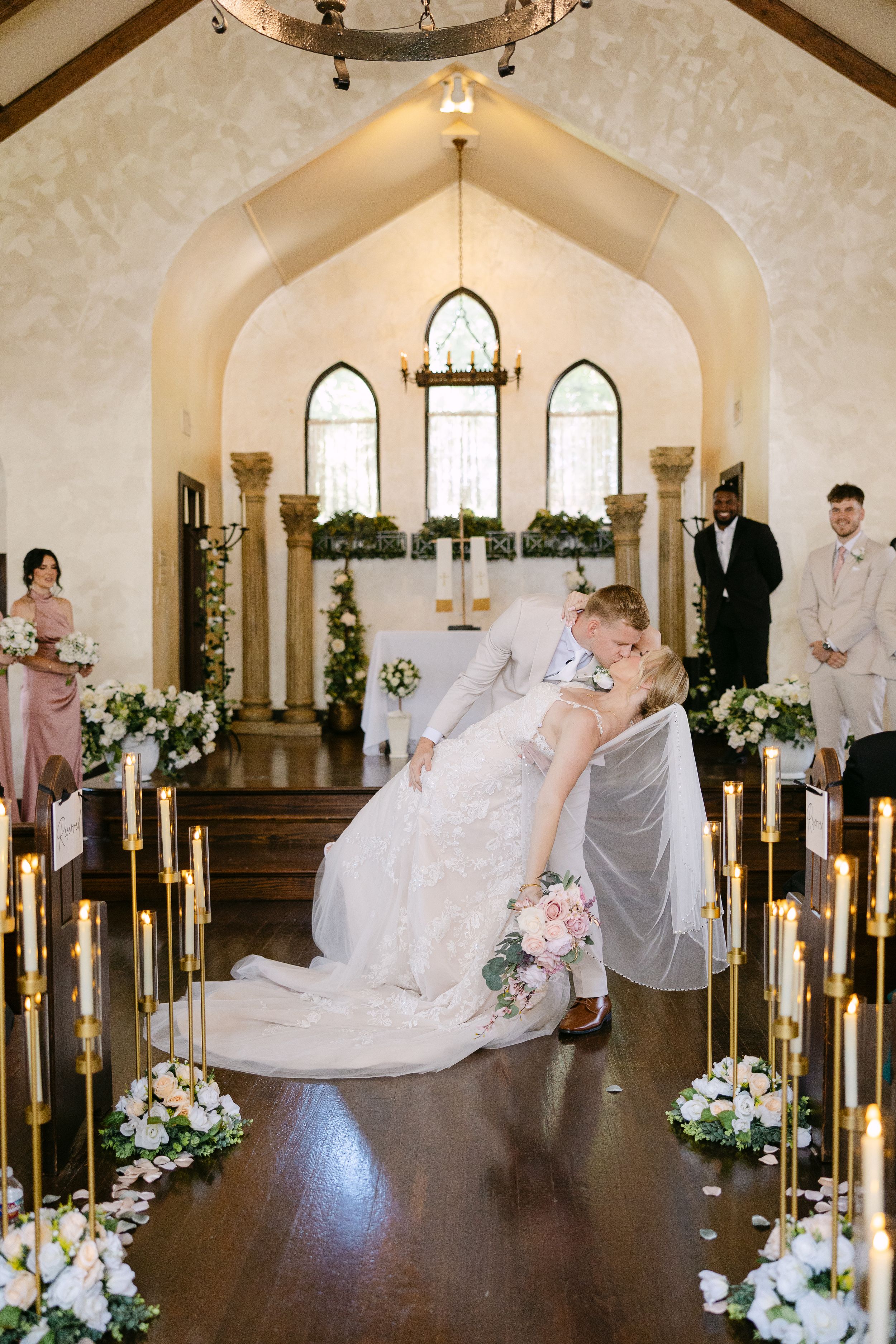 Bride and groom share a romantic dip kiss at the altar of an elegant white chapel adorned with candles and floral arrangements.