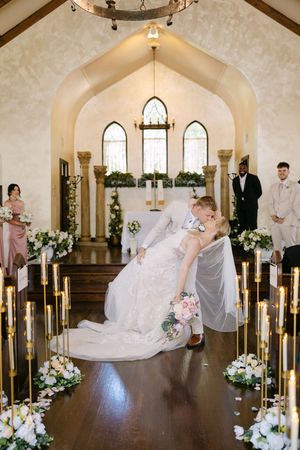 Bride and groom share a romantic dip kiss at the altar of an elegant white chapel adorned with candles and floral arrangements.