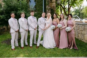 Wedding party poses outdoors, bride in white gown flanked by groomsmen in tan suits and bridesmaids in dusty rose dresses.