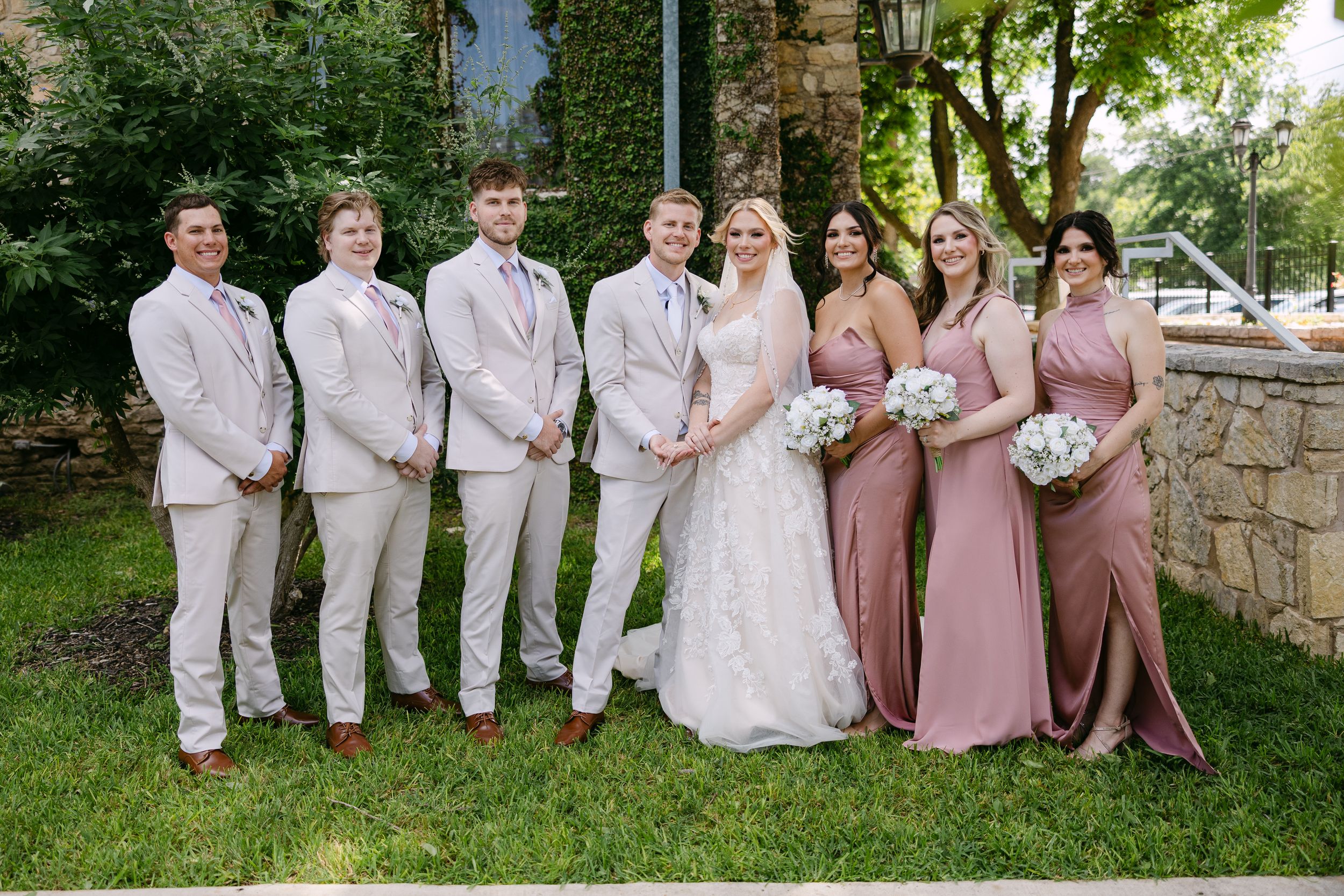 Wedding party poses outdoors, bride in white gown flanked by groomsmen in tan suits and bridesmaids in dusty rose dresses.