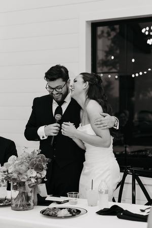 Black and white photo of a bride and groom sharing a microphone and laughing together at their wedding reception.
