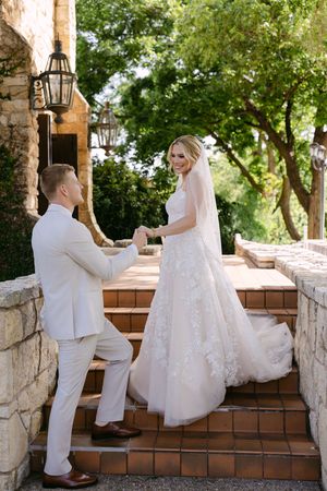 Bride and groom hold hands on terracotta steps at an outdoor wedding venue surrounded by lush greenery.