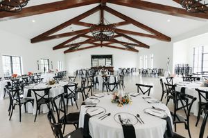 Elegant wedding reception hall with white round tables, black cross-back chairs, and rustic wood beam cathedral ceiling.