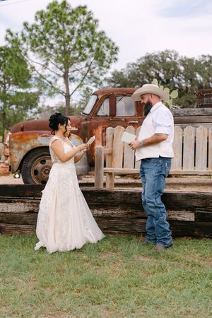 Bride and groom exchange vows outdoors at rustic country wedding with vintage truck backdrop.