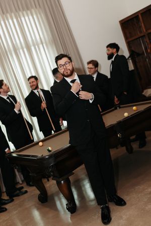 Men in black suits pose near pool tables in an elegant room, with one man adjusting his tie in the foreground.