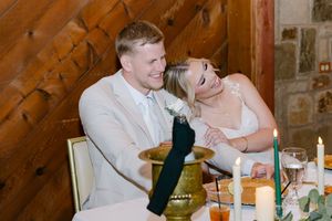 Bride and groom laughing together at wedding reception table with candles and rustic wood backdrop.