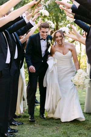 Bride and groom walk through an arch of raised arms formed by wedding party on green lawn.