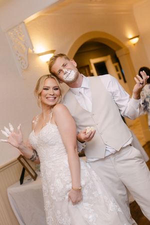 Bride and groom laugh with wedding cake on their faces during fun reception cake cutting celebration.