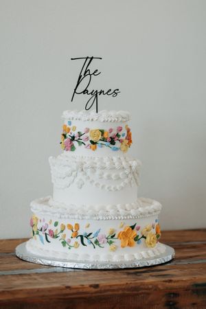 Three-tier white wedding cake with colorful floral decorations and 'The Raynes' cake topper on rustic wood table.