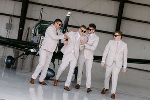 Four groomsmen in tan suits and sunglasses pose playfully in an airplane hangar with a helicopter behind them.