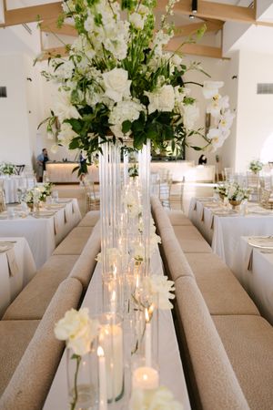 Elegant wedding reception with tall white floral centerpieces, candles, and gold chairs in a bright venue.
