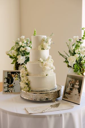 Elegant four-tier white wedding cake adorned with roses on a silver stand, flanked by floral arrangements and framed photos.