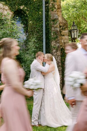 Bride and groom share a kiss while wedding party walks past in blurred motion at an ivy-covered venue.