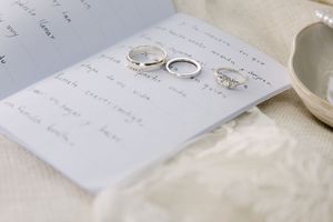 Wedding rings and diamond engagement ring resting on handwritten vows card with elegant white fabric background.