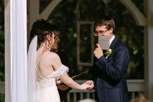 Groom reads wedding vows from a booklet while holding hands with bride in white lace dress at outdoor ceremony.