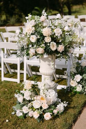 Elegant outdoor wedding ceremony with white floral arrangements in classic urns, featuring cream roses and greenery on lush grass.