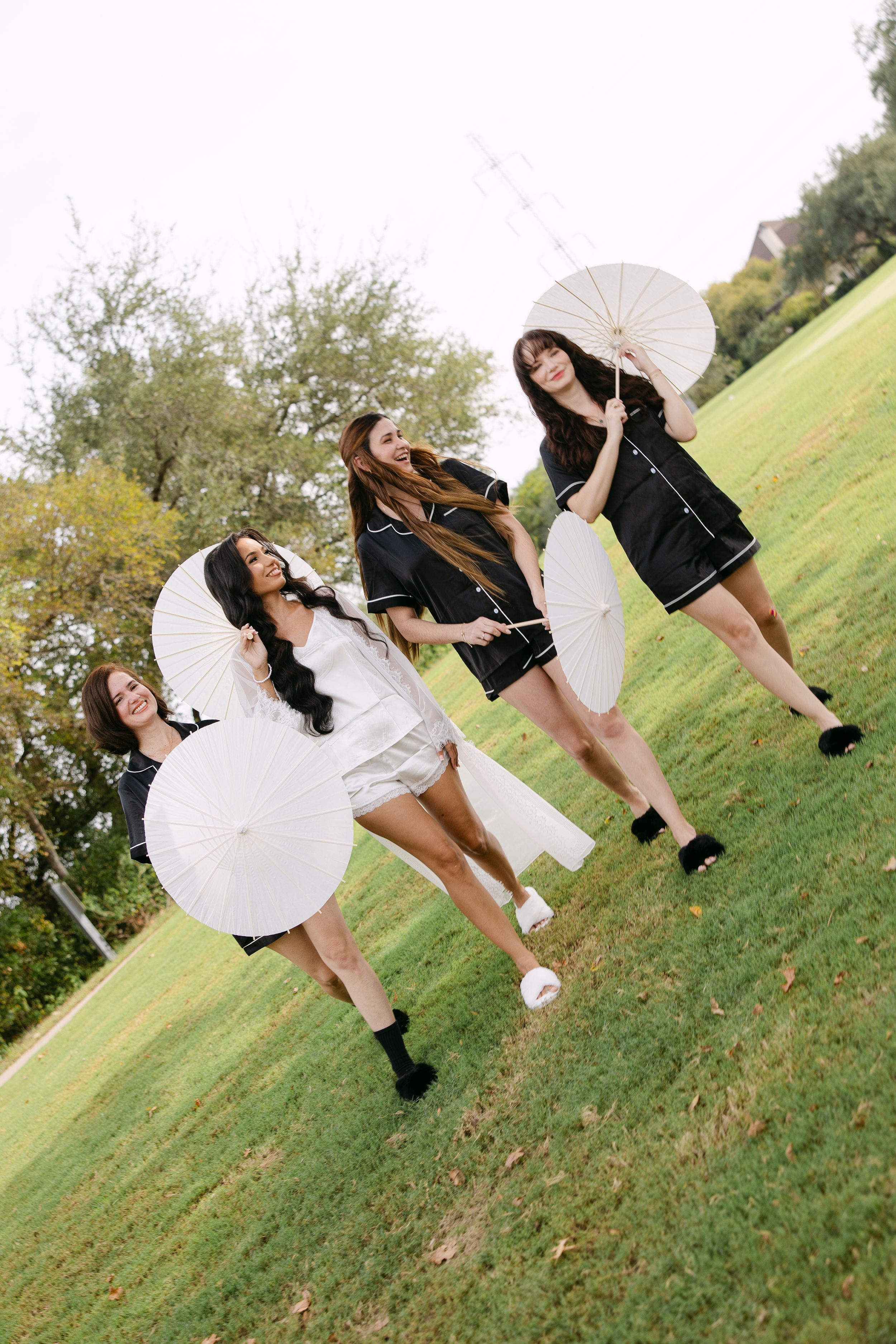Four women in black and white pajamas hold white parasols while posing playfully on a green lawn outdoors.