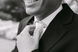 Black and white photo of a smiling groom in a suit adjusting his tie on his wedding day.