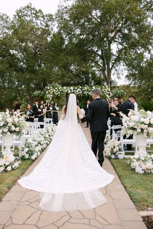 Bride walking down outdoor aisle with escort, white floral arch and arrangements, guests seated in white chairs.