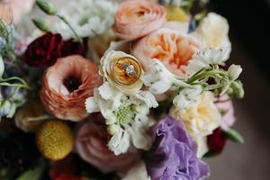 Close-up of wedding rings resting on a colorful bridal bouquet with roses, ranunculus, and purple hydrangeas.