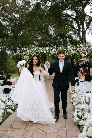 Bride and groom walk down outdoor wedding aisle lined with white floral arrangements, holding hands and smiling joyfully.