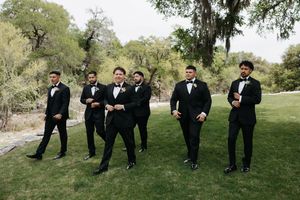 Six groomsmen in black tuxedos with bow ties pose casually on green lawn surrounded by lush trees at outdoor wedding.