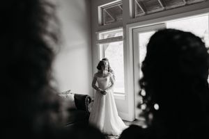 Black and white photo of a bride in a white gown standing by a window, viewed between two silhouetted figures.