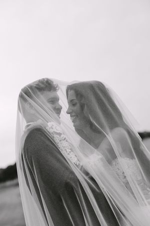 Black and white photo of bride and groom smiling under wedding veil.