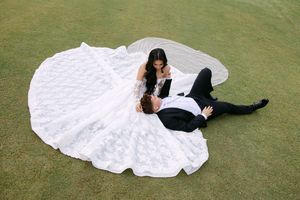 Bride in white lace gown and groom in tuxedo pose romantically on green grass, aerial wedding photo.