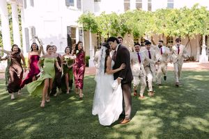 Bride and groom share a romantic kiss surrounded by bridesmaids in colorful dresses and groomsmen on a lush green lawn.
