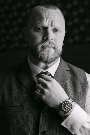 Black and white portrait of a bearded bald man in a suit adjusting his tie, wearing a watch.