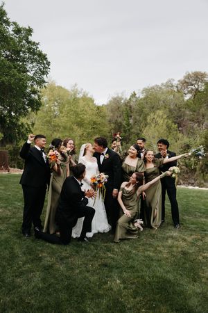 Wedding party celebrates as bride and groom kiss outdoors, bridesmaids in sage green gowns and groomsmen in black tuxedos.
