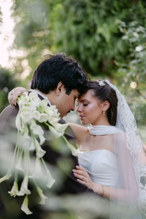Bride and groom share an intimate forehead touch moment, surrounded by lush greenery, bride holding white calla lily bouquet.