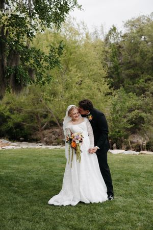 Bride in white lace gown holding colorful bouquet as groom in black suit kisses her cheek on green lawn.