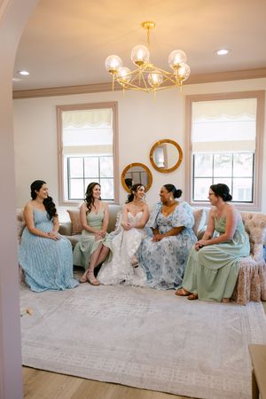 Bride and four bridesmaids in sage green dresses sit together in an elegant bridal suite with gold chandelier.