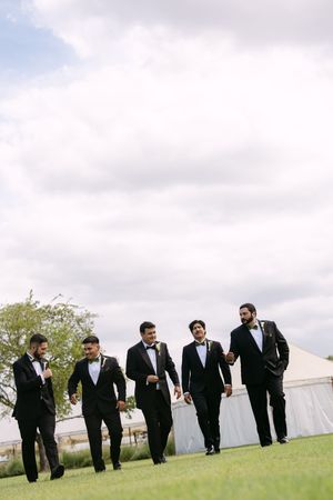 Five groomsmen in black tuxedos and bow ties walk across green lawn outdoors under cloudy sky at wedding venue.