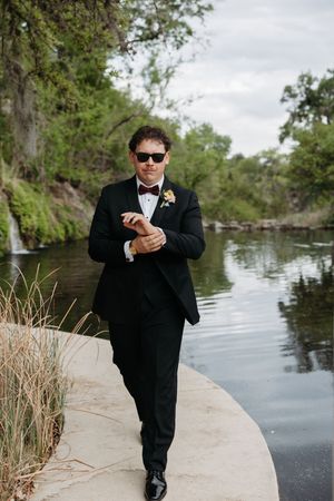 Groom in black tuxedo with bow tie and sunglasses adjusting cufflinks beside a serene river surrounded by lush greenery.