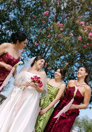 Bride in white gown with three bridesmaids in red and green dresses, laughing outdoors under blooming pink trees.
