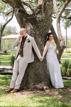 Bride and groom hold hands leaning against a large oak tree, groom wiping tears in a tan suit, bride in a lace gown.