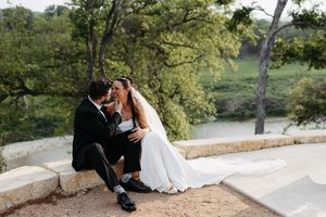 Bride and groom sitting together on a stone ledge, sharing a romantic moment with lush green trees in the background.
