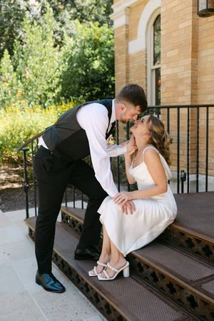 A groom in a black suit leans down to kiss his bride in a white dress seated on ornate steps outside a brick building.