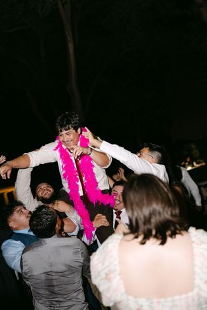Groom lifted by guests wearing pink feather boa at outdoor wedding reception celebration at night.