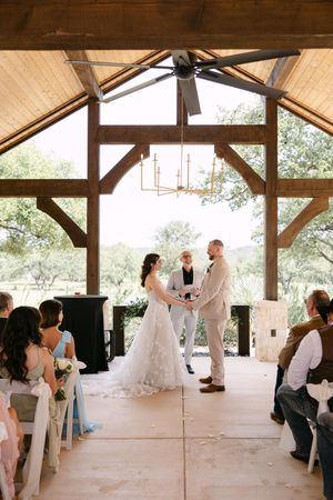 Bride and groom exchange vows at a rustic outdoor wedding chapel with wooden beams and lush greenery backdrop.