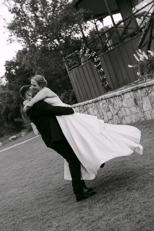 Groom lifting bride in white gown outdoors at wedding venue, black and white romantic portrait.