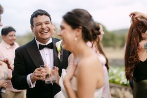 Groom in tuxedo laughing joyfully with bride in white dress during outdoor wedding reception celebration.