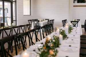 Elegant wedding reception with long tables, black cross-back chairs, candles, and greenery garlands in a bright white venue.